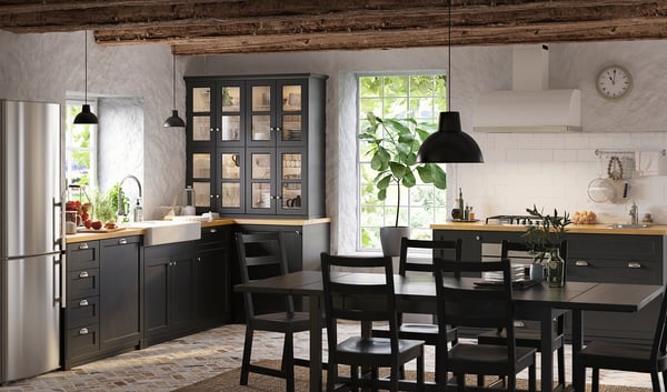 Современная кухня со шкафами из черного окрашенного ясеня, белой плиткой, холодильником из нержавеющей стали, деревянным обеденным столом, подвесными светильниками и растением в горшке.