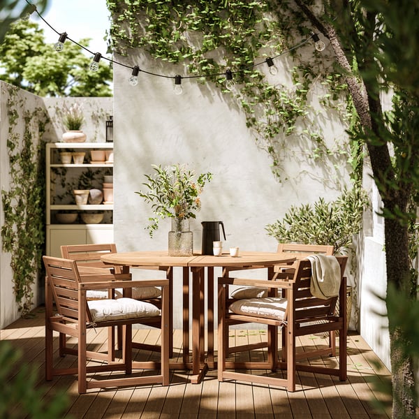 Обеденный гарнитур на открытом воздухе с деревянным столом и мягкими стульями, украшенными гирляндами наверху, на фоне белой стены.