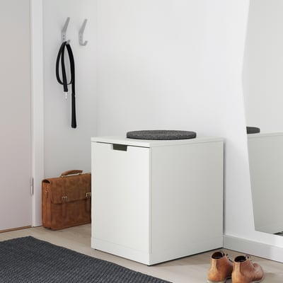 Современный белый сундук NORDLI с серой подушкой, рядом со входом с приветственным ковриком, сумкой и полкой для обуви.