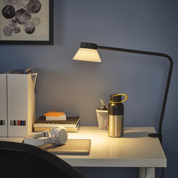 Настольный светильник SAMBORD с треугольным абажуром, освещающий рабочее место с книгами, бутылкой и наушниками.