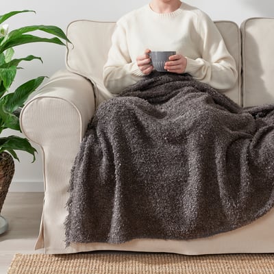 Человек, сидящий на бежевом диване с кружкой в руках, накрытый уютным серым одеялом SANDBRODD с бахромой.
