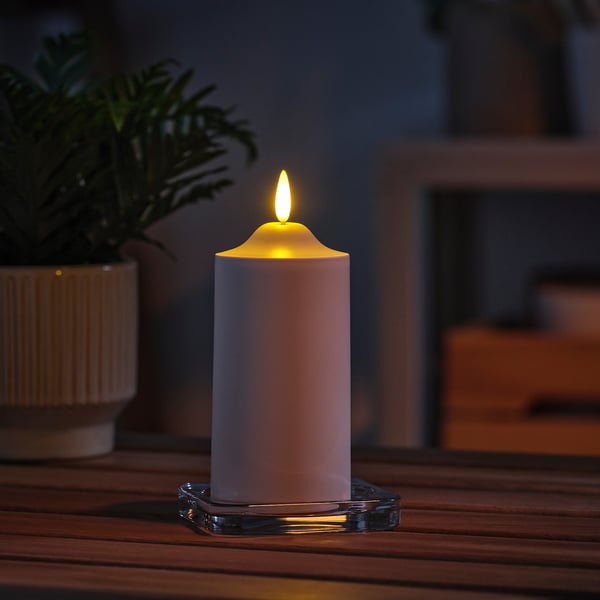 Белая светодиодная свеча Ikea SMÅLOM на подстаканнике, тепло светящаяся на деревянном столе рядом с зеленым растением.