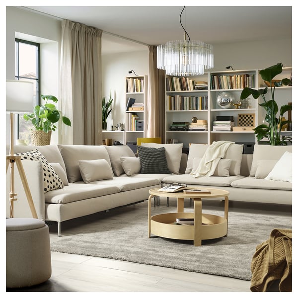 Современная гостиная с бежевым диваном SÖDERHAMN, подушками, деревянным журнальным столиком, книжной полкой, растениями, ковром и люстрой.