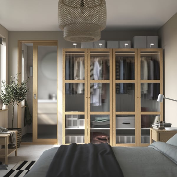 Большая спальня с деревянным шкафом, стеклянными дверями, зеркалом, кроватью, тумбочкой, лампой, растением и плетеным светильником.