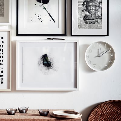 Настенные художественные рамки и белые часы. На циферблате часов цифры отображаются в виде точек, черные стрелки показывают время. Внизу на плетеной корзине и столе стоят небольшие пиалы.