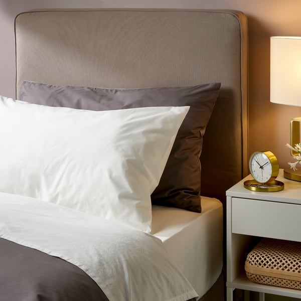Спальня с серой наволочкой ULLVIDE на белой кровати. Нейтральные цвета, гладкие простыни, красиво оформленная прикроватная тумбочка с часами и лампой.