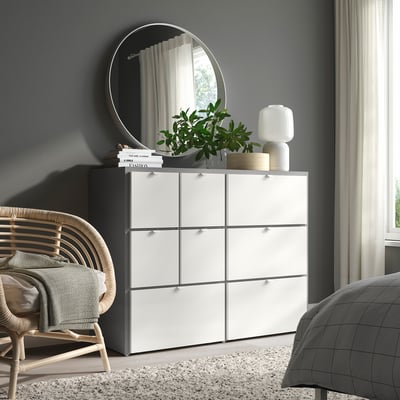 Современная спальня с комодом, зеркалом, светильником, растением, плетеным креслом, серой кроватью, окном.