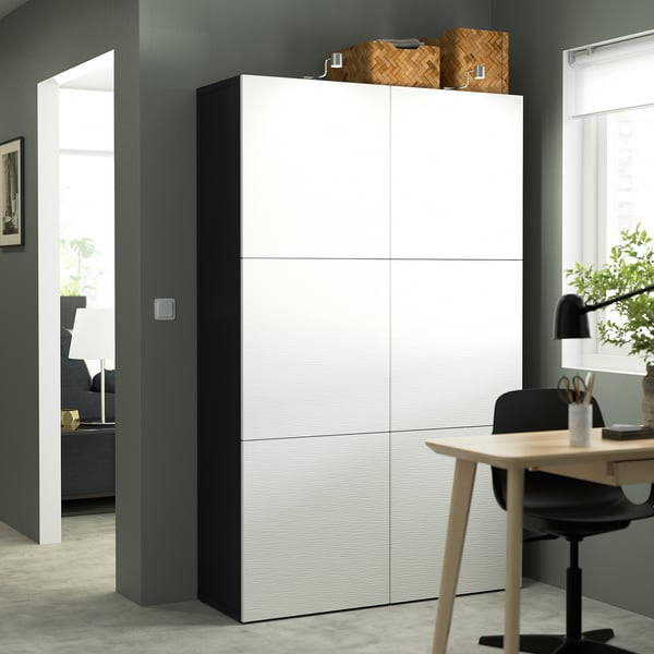 Large white BESTÅ storage cabinet against dark wall, taller than nearby desk and chair.