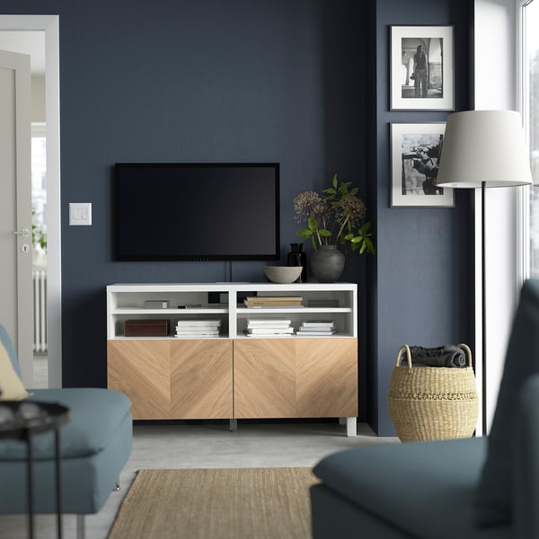 White BESTÅ tv bench, flat screen tv, blue chairs, basket, lamp, dark walls, photos, couch, rug.