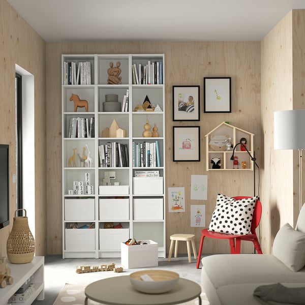 Modern living room with BILLY white bookcases, red chair, polka dot pillow, wooden walls, and various childrens toys and books.