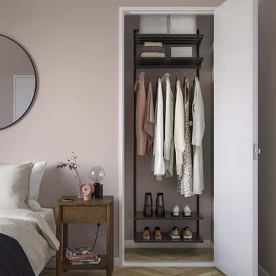 BOAXEL closet storage with clothes and accessories. Steel, wall-mounted, dark grey. Nearby bedside table with lamp, clock, plant.