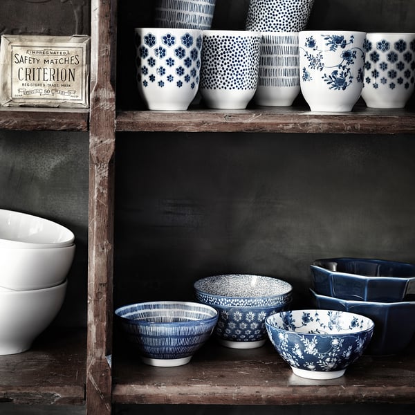 Collection of blue and white ceramic mugs and bowls with floral and geometric designs, arranged on rustic wooden shelves.