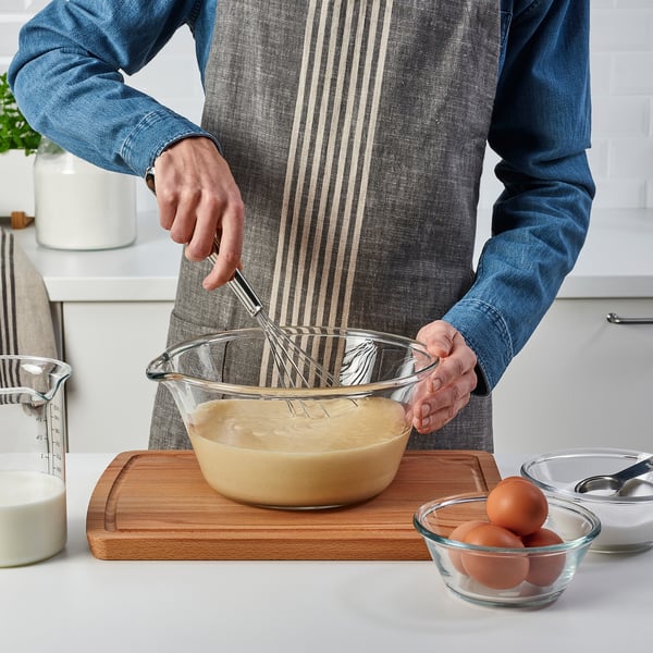 Person mixing batter with FINMAT whisk in glass bowl on cutting board, surrounded by ingredients.