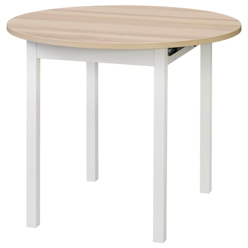 Depicts a round, extendable wooden dining table with white legs. Its designed for 2-4 people and has a smooth, light-colored top.