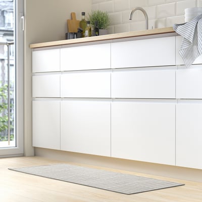 Modern kitchen with white cabinets. Grey & white striped mat on floor, fitting under sink. Wooden countertop with green plant, bottles, and towels.