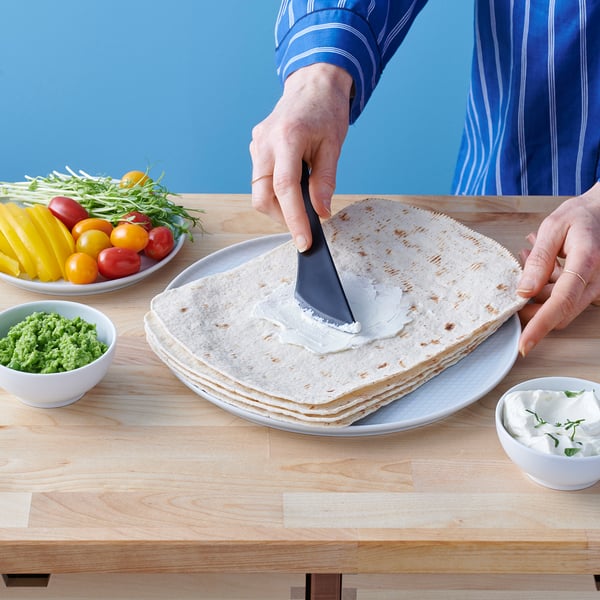 Person spreading cream on flatbread with knife. Colourful veggies and herbs nearby.