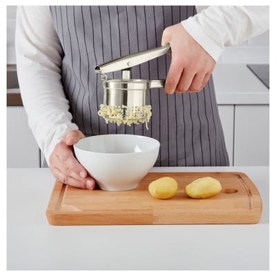 Person presses potatoes through a stainless steel potato press over a bowl on a wooden cutting board for quick mashing.