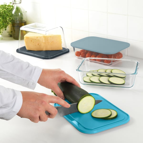 Person slices courgette on blue chopping board/lid. Nearby, cheese and tomatoes stored in IKEA 365+ containers.