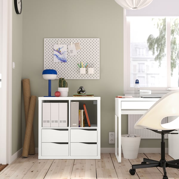 KALLAX shelving with books, plant, lamp, and notes. White desk with computer and chair near window.