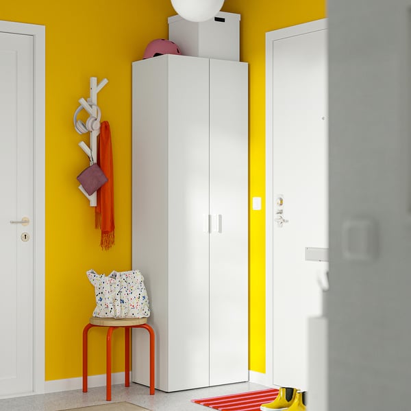 Compact white LASTARE wardrobe in vibrant yellow room, with red stool, pre-drilled holes, topped with boxes and a lamp.