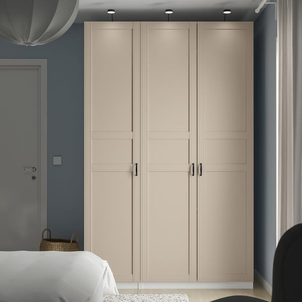 Beige PAX wardrobe against blue wall, flanked by bed and chair. Tall, sleek handles, subtle panelling.