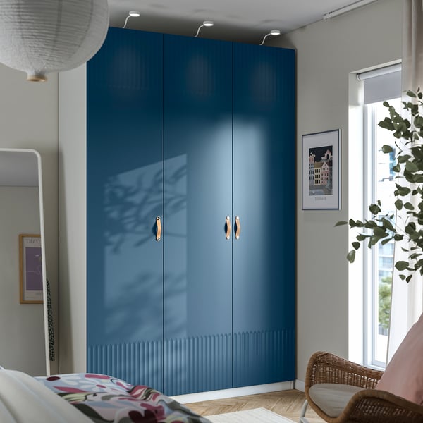 A large blue PAX wardrobe with gold handles, situated between a bed and a window with a plant.