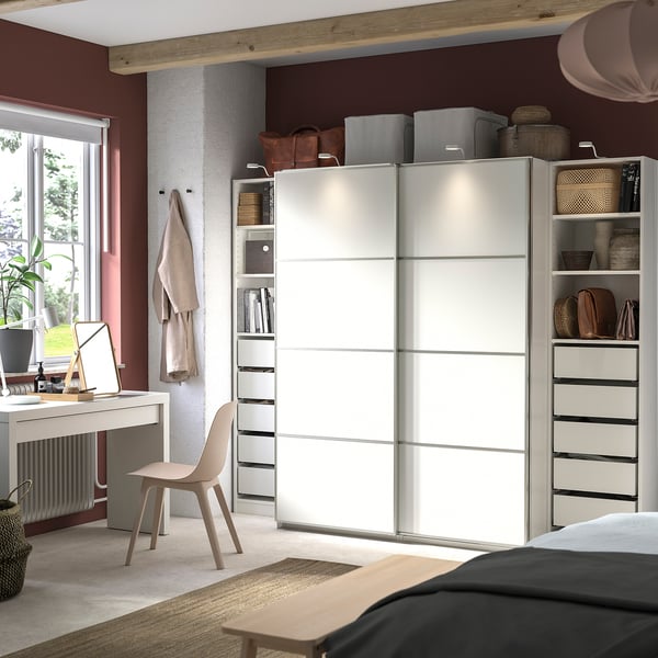 Modern bedroom with a white PAX wardrobe featuring sliding doors and shelves. Includes a desk, chair, and natural decor elements.