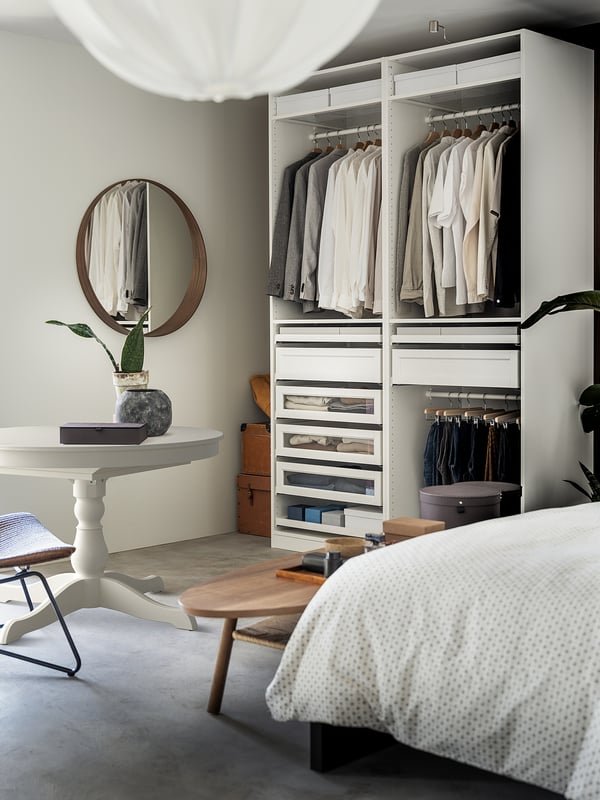 Modern bedroom with white PAX wardrobe, round mirror, white bed, and wooden table. Clothes organised neatly, grey and white.
