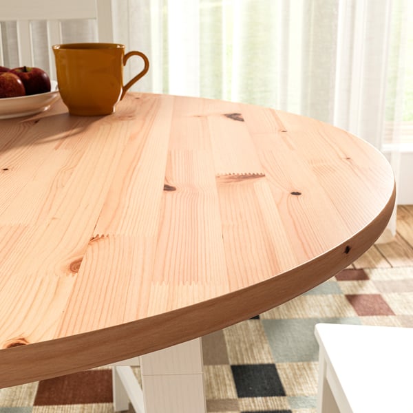 Light wood round table with smooth edges. Mug and fruit on top, placed near window. Bright, natural light fills room.