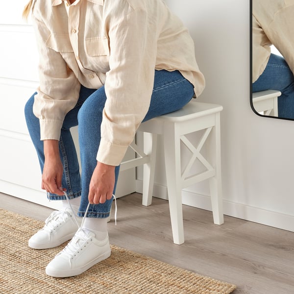 Person tying white shoelaces, sitting on white ROSENTORP stool, with beige shirt and jeans.