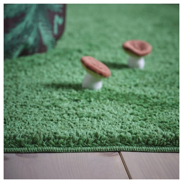 Soft, plush green rug mimics forest grass. Two toy mushrooms sit atop, texture vivid.