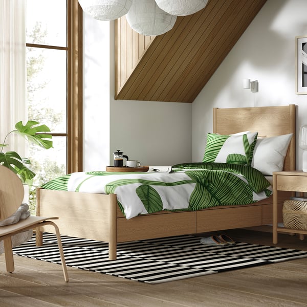 Modern bedroom with TONSTAD bed, leaf-pattern bedding, drawers, large window, sheer curtains, rug, chair, plant. Airy, natural light.