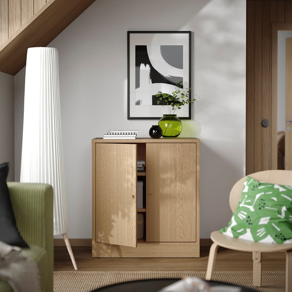 Wooden cabinet between lamp & chair, with vase, candle, books. Abstract art above, silent soft-close, adjustable shelves.