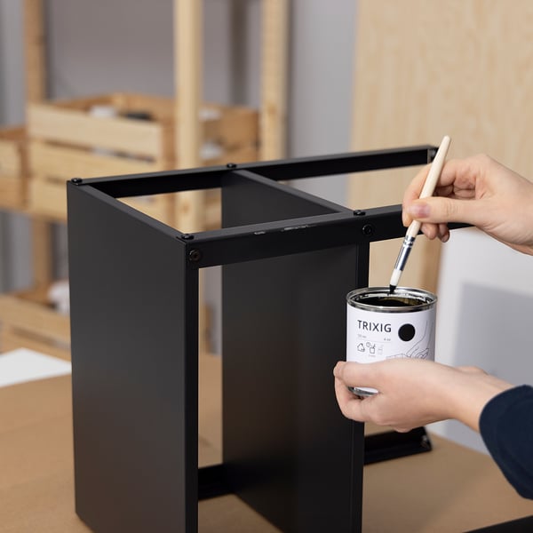 A person is painting a black metal frame with a can labelled TRIXIG.