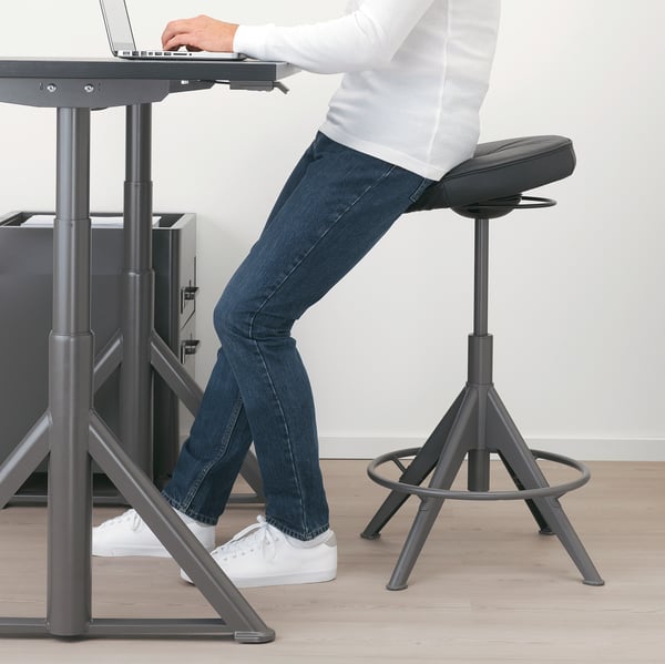 Person sitting on adjustable stool, typing on laptop at high desk.
