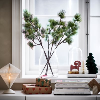 VINTERFINT, green pine stem in clear vase, decorated window sill, surrounded by books, gifts, and festive decor.
