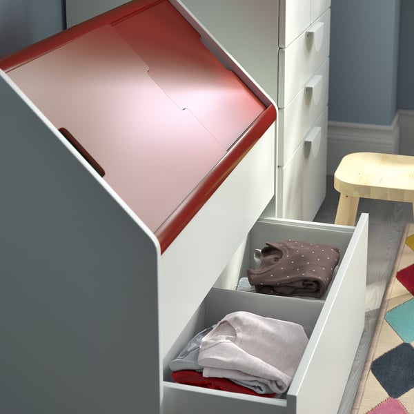 Open white VITBJÖRN storage unit with grey drawers, one holding folded grey and red clothes, against blue wall.