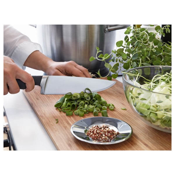 Person chopping scallions on a wooden cutting board with a stainless steel knife. A small plate of chilli flakes is nearby.
