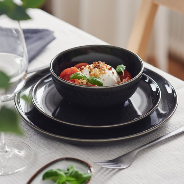 Темная миска на сложенных друг на друга тарелках, подача моцареллы, помидоров и базилика. Элегантная сервировка стола с бокалами и столовыми приборами.