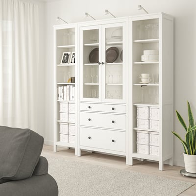 Белый шкаф HEMNES со стеклянными дверцами, полками и ящиками рядом с серым диваном, где выставлена стеклянная посуда, посуда и книги.