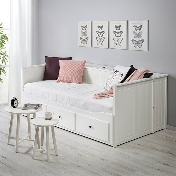 HEMNES белая кушетка с местом для хранения, подушками и одеялом. Небольшие приставные столики с кружкой и книгой. Принты с бабочками на стенах.