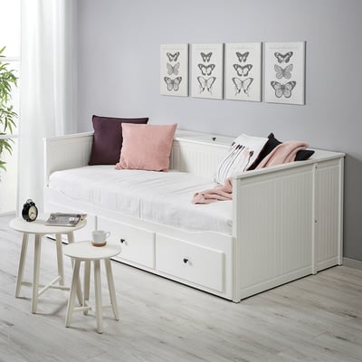 HEMNES белая кушетка с местом для хранения, подушками и одеялом. Небольшие приставные столики с кружкой и книгой. Принты с бабочками на стенах.