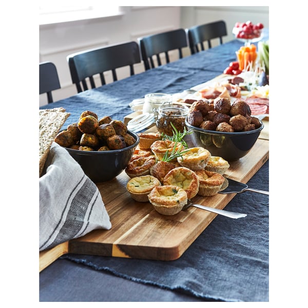 Черные миски с едой на деревянной разделочной доске, поставленные на серый стол с черными стульями вокруг.