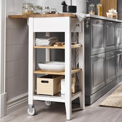 Белая кухонная тележка STENSTORP с деревянными полками. Хранит банки, полотенца и миски. Рядом шкафы и кафельная стена.