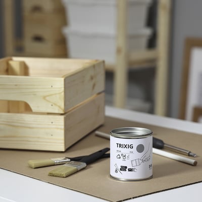 Маленькая баночка серой краски TRIXIG лежит на картоне, окруженная кистями, отверткой и деревянной коробкой.