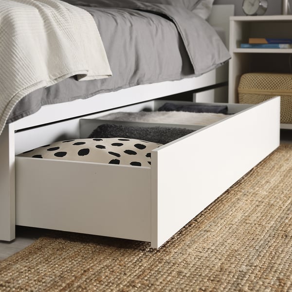 Ящики для хранения под кроватью с узорчатыми вкладышами раскрываются для удобной организации. Ящики расположены под простым белым каркасом кровати.
