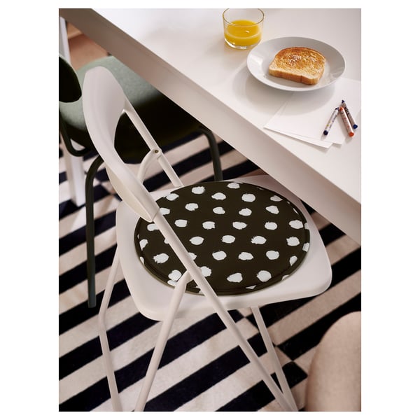 Image ÄNGSFRÄKEN chair pad on white chair by dining table. Pad is black with white polka dots, round shape, made from recycled polyester.