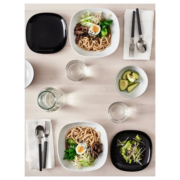 A minimalist table setting with square, black, tempered glass plates and bowls, noodle dishes, avocado, and utensils.