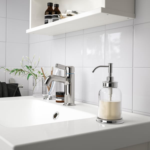 Modern chrome bathroom sink soap dispenser sits right beside tap, smaller than the glass vase with green plant.