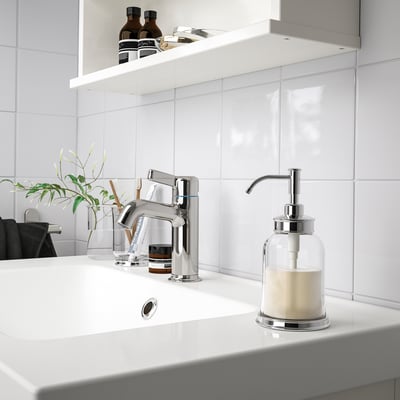 Modern chrome bathroom sink soap dispenser sits right beside tap, smaller than the glass vase with green plant.